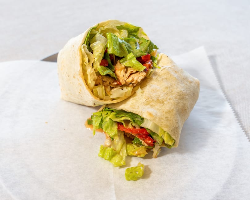 A chicken wrap, cut in half, filled with lettuce, red peppers, and chicken, on white paper.