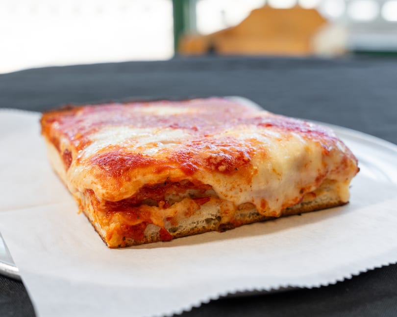 Thick square slice of Detroit-style pizza with melted cheese and red sauce on parchment paper.