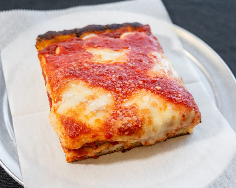 A thick square slice of Detroit-style pizza with melted cheese and red sauce on parchment paper.