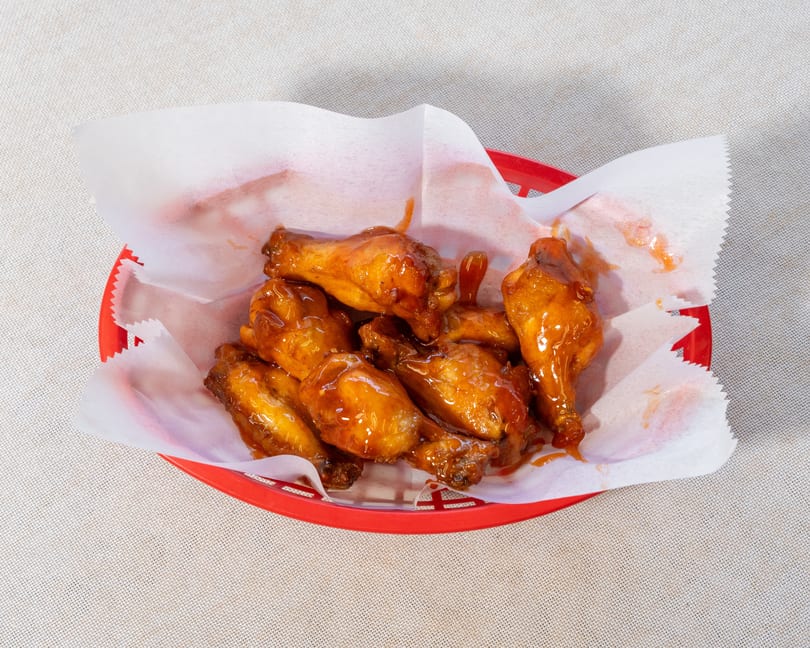 A red plastic basket filled with several glistening, saucy chicken wings on white parchment paper.