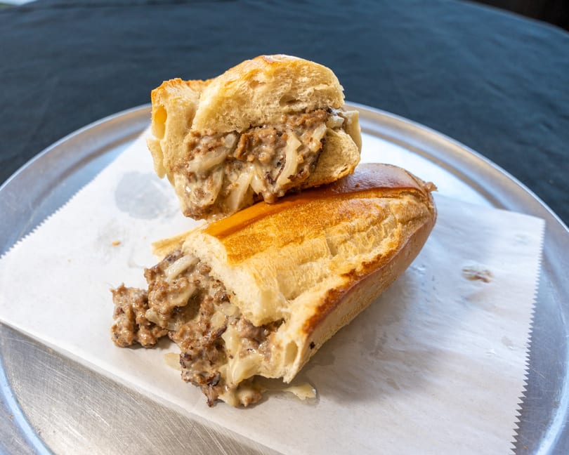 A cheesesteak sandwich, cut in half, filled with meat, onions, and melted cheese on a metal tray.