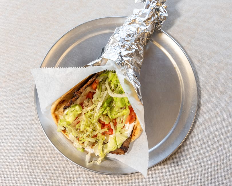 A gyro with meat, lettuce, tomatoes, and sauce, partially wrapped in foil on a metal plate.