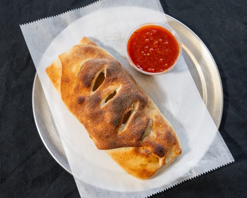 Golden-brown calzone with slits on top, served with a side of red marinara sauce on a metal plate.
