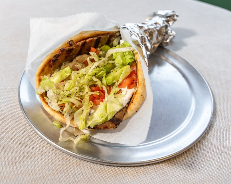A gyro with meat, lettuce, tomatoes, and white sauce, partially wrapped in foil on a metal plate.
