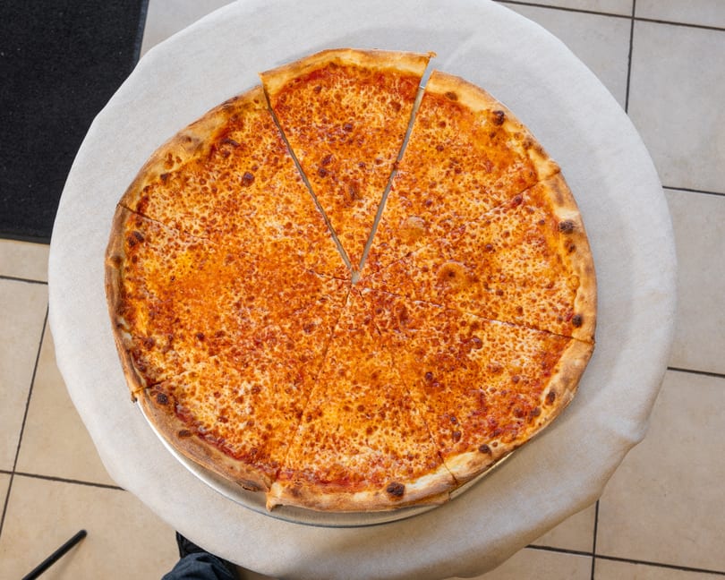 A large, round cheese pizza with two slices removed, on a metal tray atop a white tablecloth.