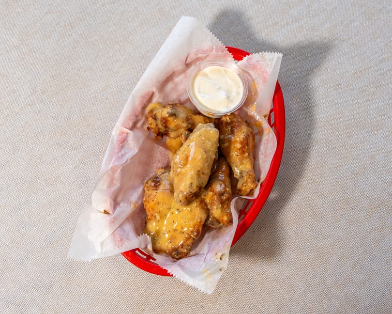 A red basket of saucy chicken wings with a side of ranch dressing on a light-colored tablecloth.
