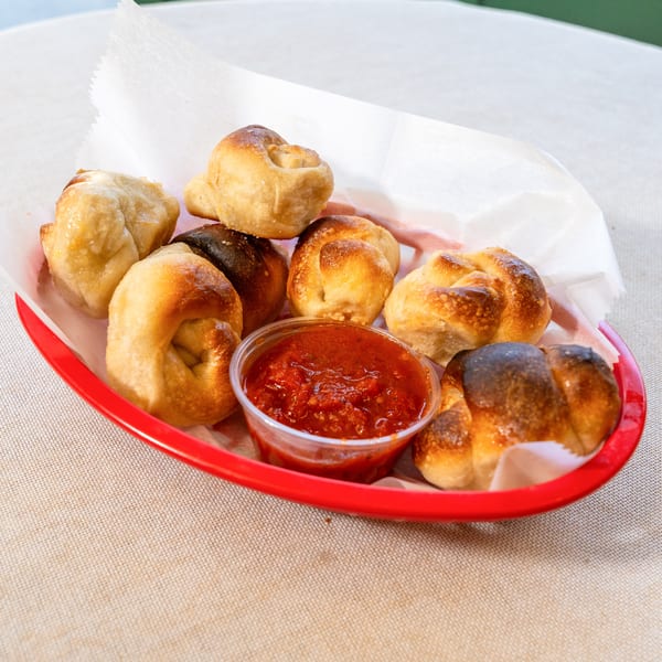 A red basket filled with golden-brown garlic knots and a small cup of marinara sauce.