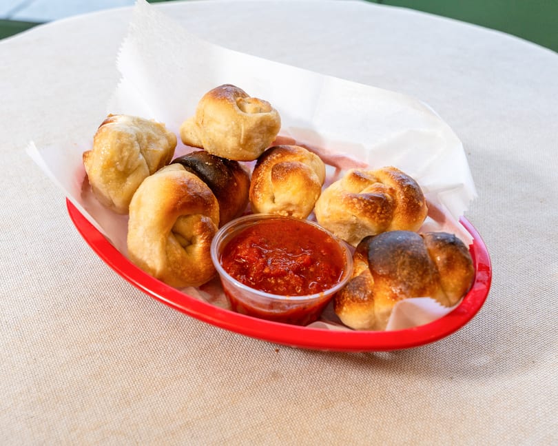 A red basket filled with golden-brown garlic knots and a small cup of marinara sauce.